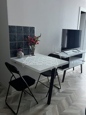 A dining area with a table, chairs, a vase with flowers, a flat-screen TV, and a herringbone-patterned floor. - Rezidenca Moderne 4R–In the Heart of Shkodër,a stylish building,central location (Shkodër)