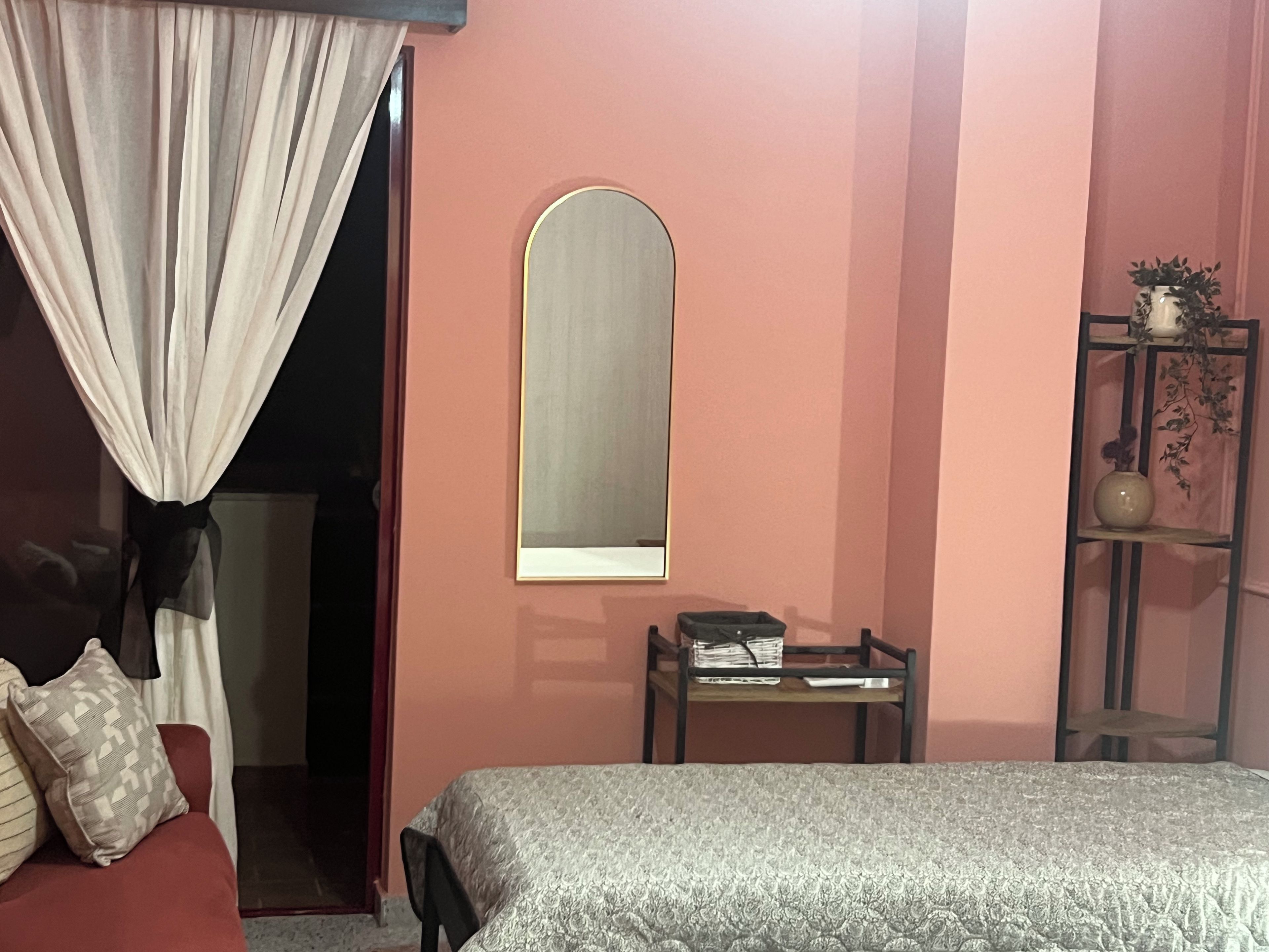 A bedroom with a bed, a bedside table, a mirror, a shelf with a vase, and a door leading to another room.