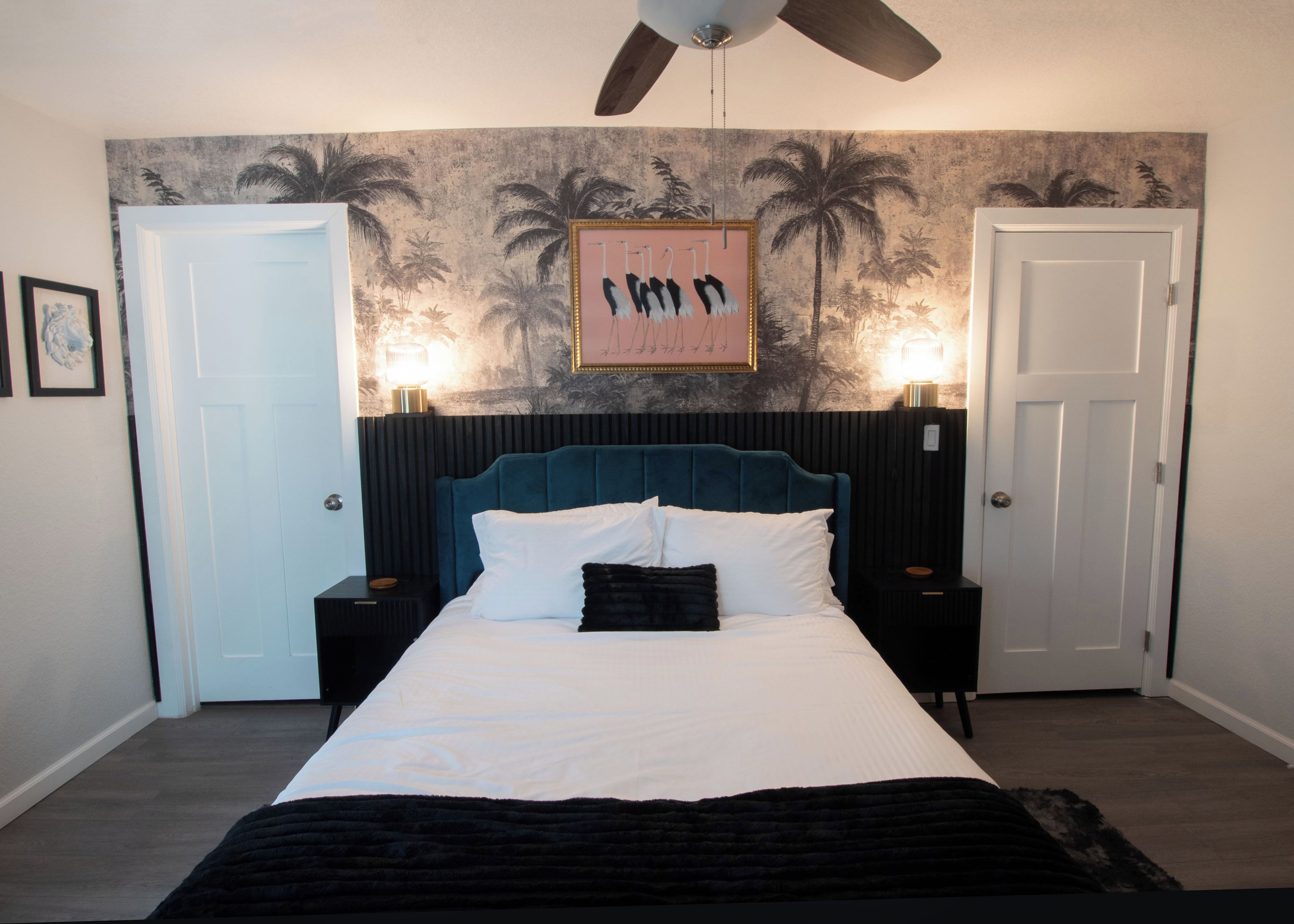 A bedroom with a bed, a ceiling fan, a wall with a tropical mural, and two bedside lamps.