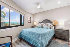 Ein Schlafzimmer mit einem großen Bett, einem Deckenventilator, Meerblick durch ein Fenster und einem Nachttisch mit Lampe.