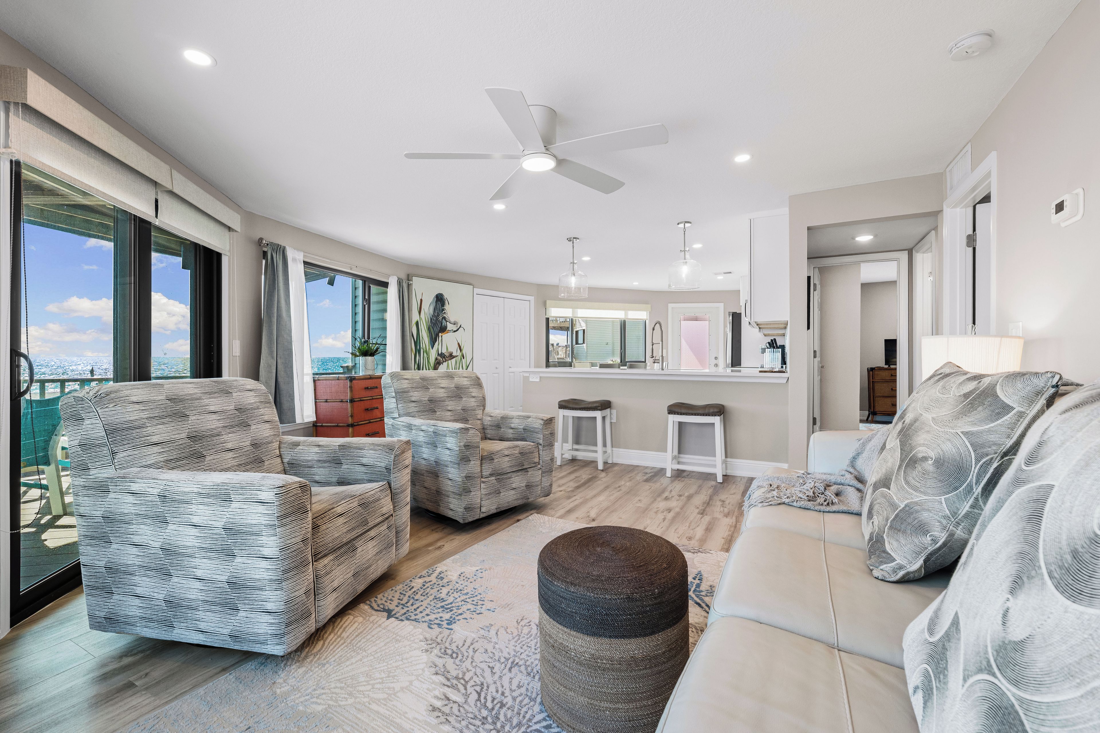 A spacious living area with a ceiling fan, a large window with a view, and a kitchen area in the background.
