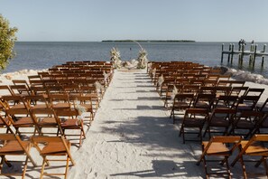 A beach wedding setup with rows of wooden chairs and a sandy aisle leading to a decorated arch.