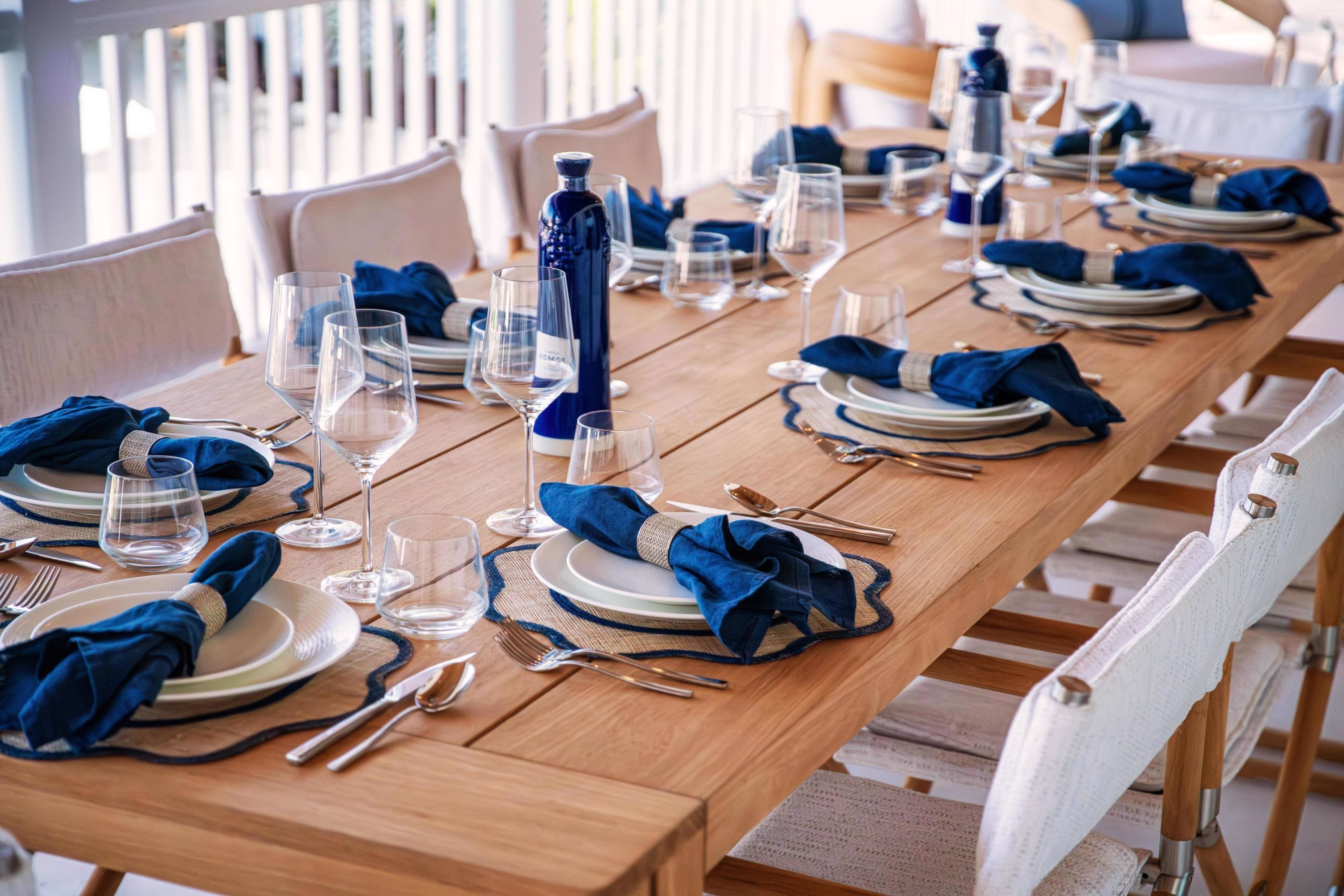 Una larga mesa de comedor dispuesta con platos, vasos y cubiertos, con servilletas azules y una botella de agua.