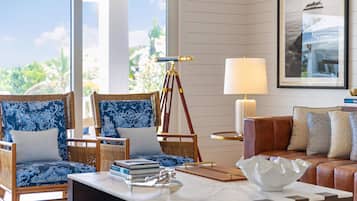 A well-lit room with a large window, two wicker chairs with blue cushions, a leather sofa, a telescope on a tripod, a table with books and a white bowl, and a framed picture on the wall.
