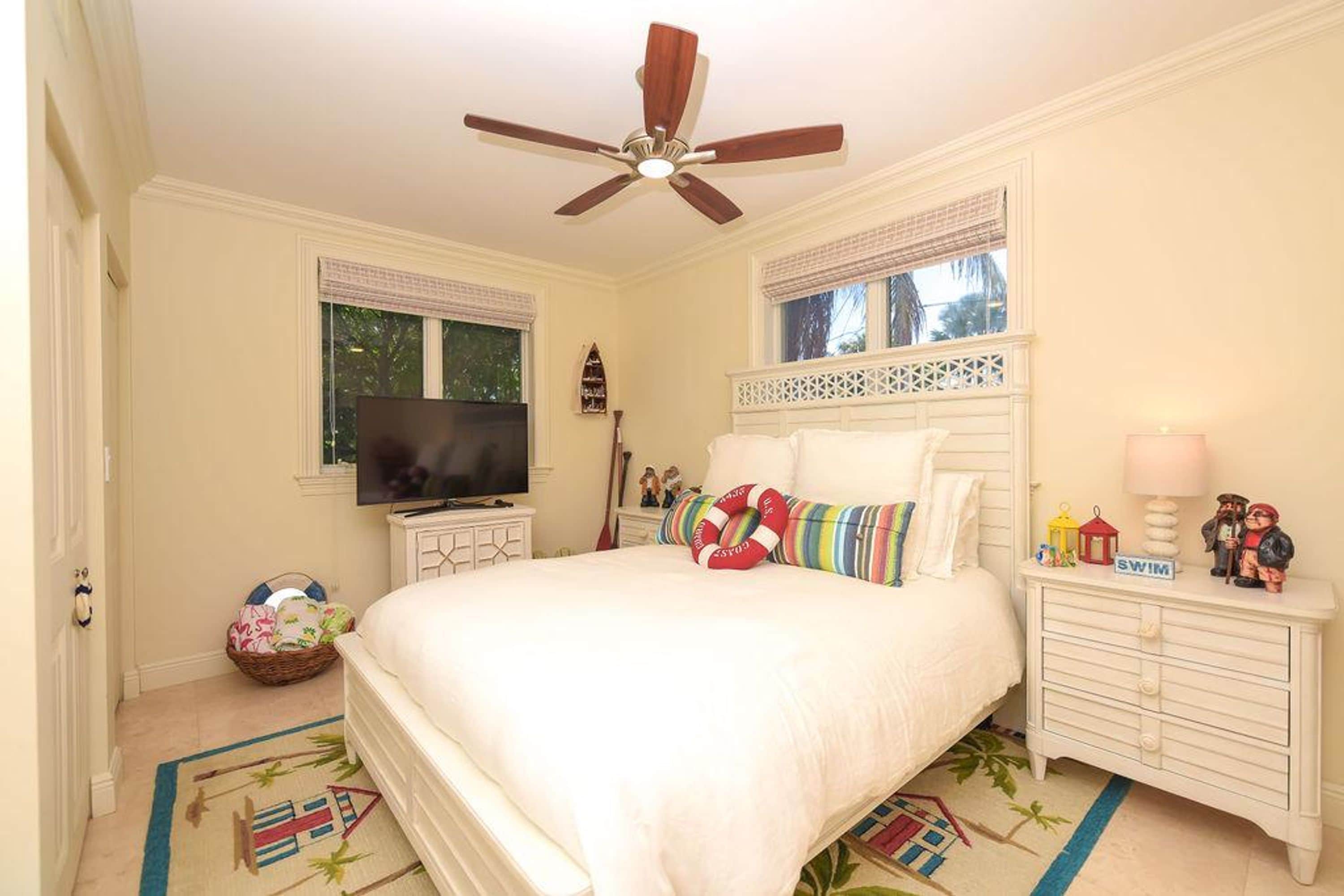 Un dormitorio con cama, televisión, ventilador de techo, mesita de noche, lámpara y ventana con persianas.