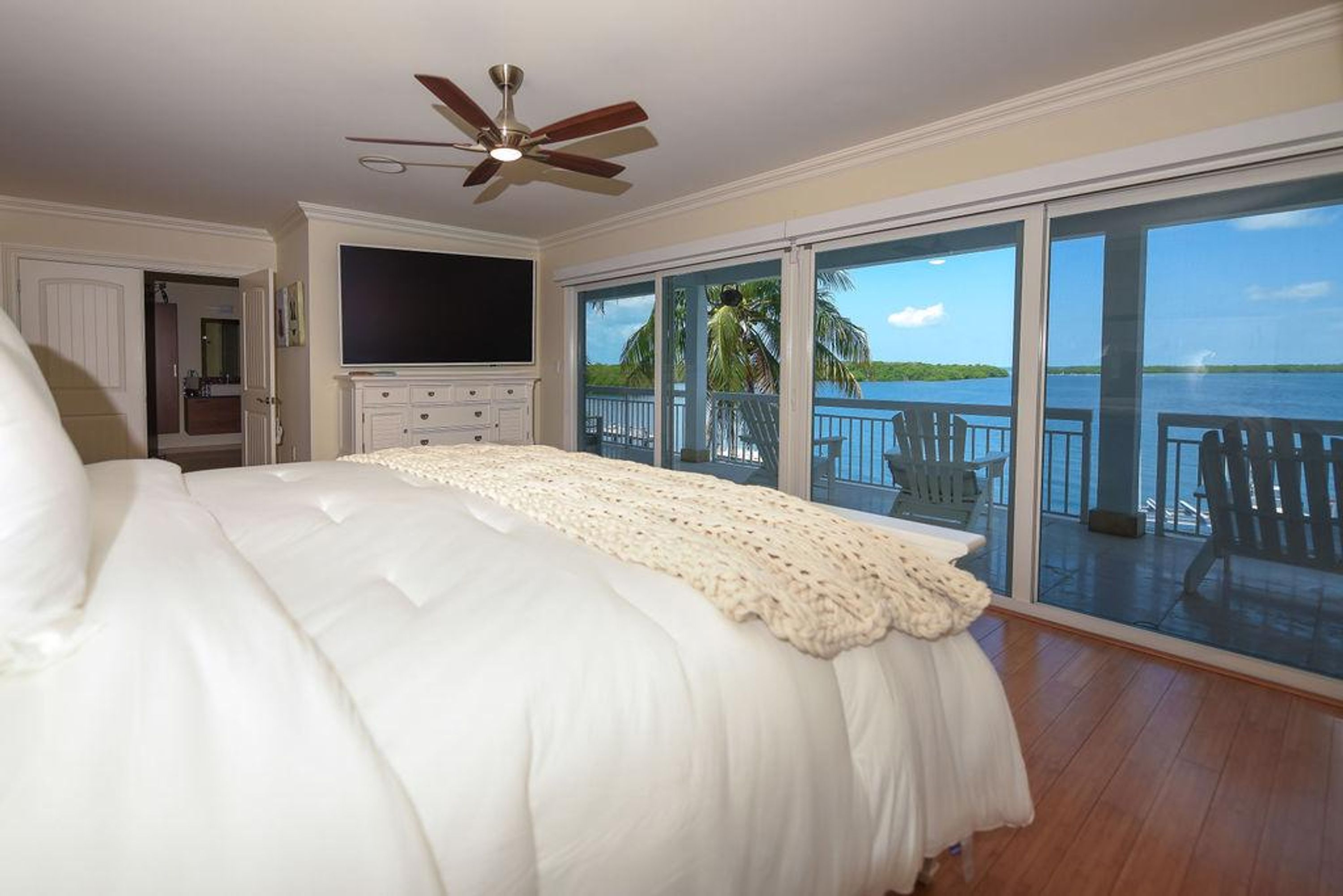 A bedroom with a large bed, a ceiling fan, a TV, and a balcony with a view of the water.
