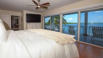 A bedroom with a large bed, a ceiling fan, a TV, and a balcony with a view of the water.