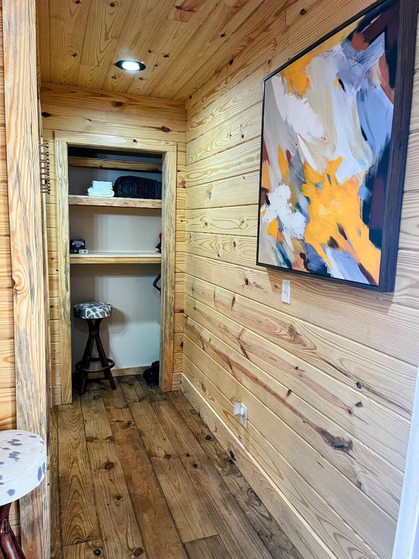 Un couloir étroit aux murs en bois et au plafond bas, qui mène à un placard doté d’étagères et d’un tabouret.