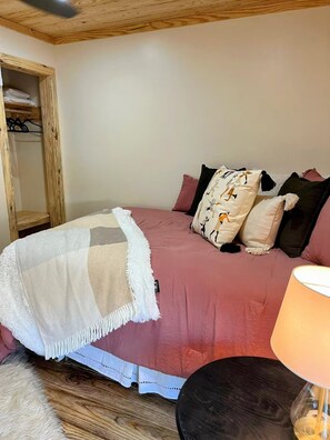 A bedroom with a bed, pillows, a blanket, a wooden headboard, a bedside table with a lamp, and a wardrobe.
