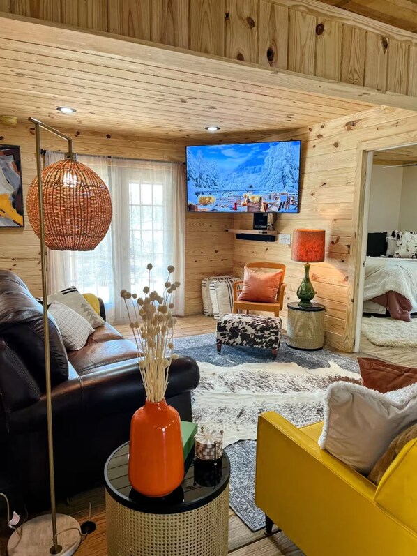 A cozy living room with a wooden ceiling, a television mounted on the wall, a black leather sofa, a yellow armchair, and a small round table with a vase.