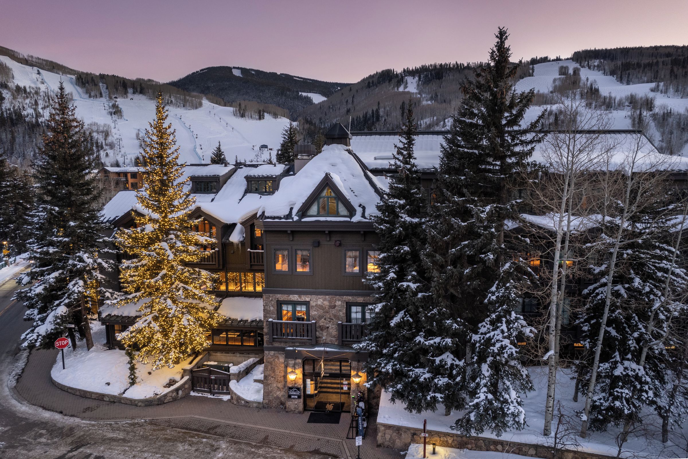 Un chalet dont le toit est recouvert de neige, doté de nombreuses fenêtres et orné d’un grand sapin de Noël illuminé.