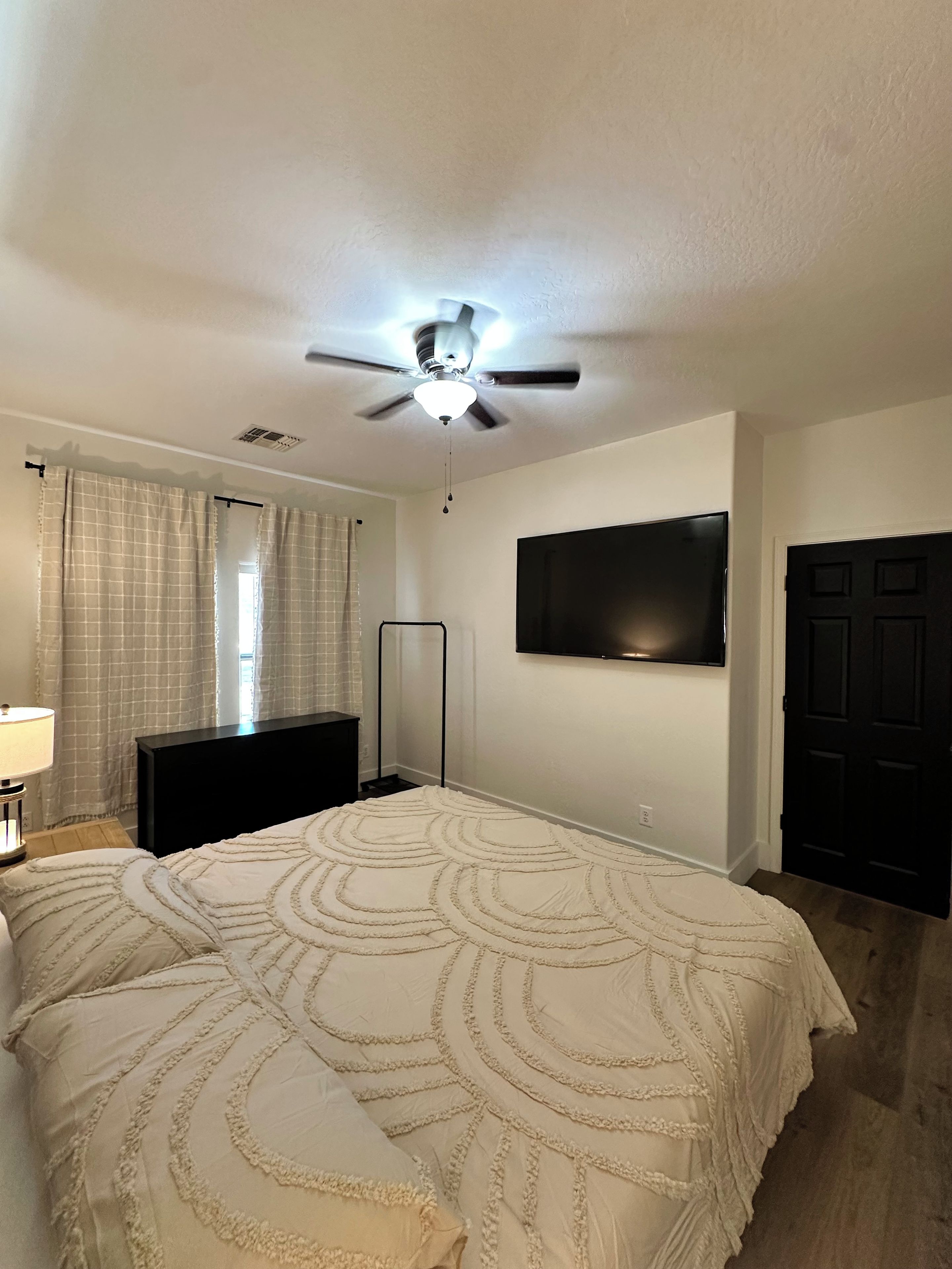 A bedroom with a bed, a ceiling fan, a TV mounted on the wall, and a door.