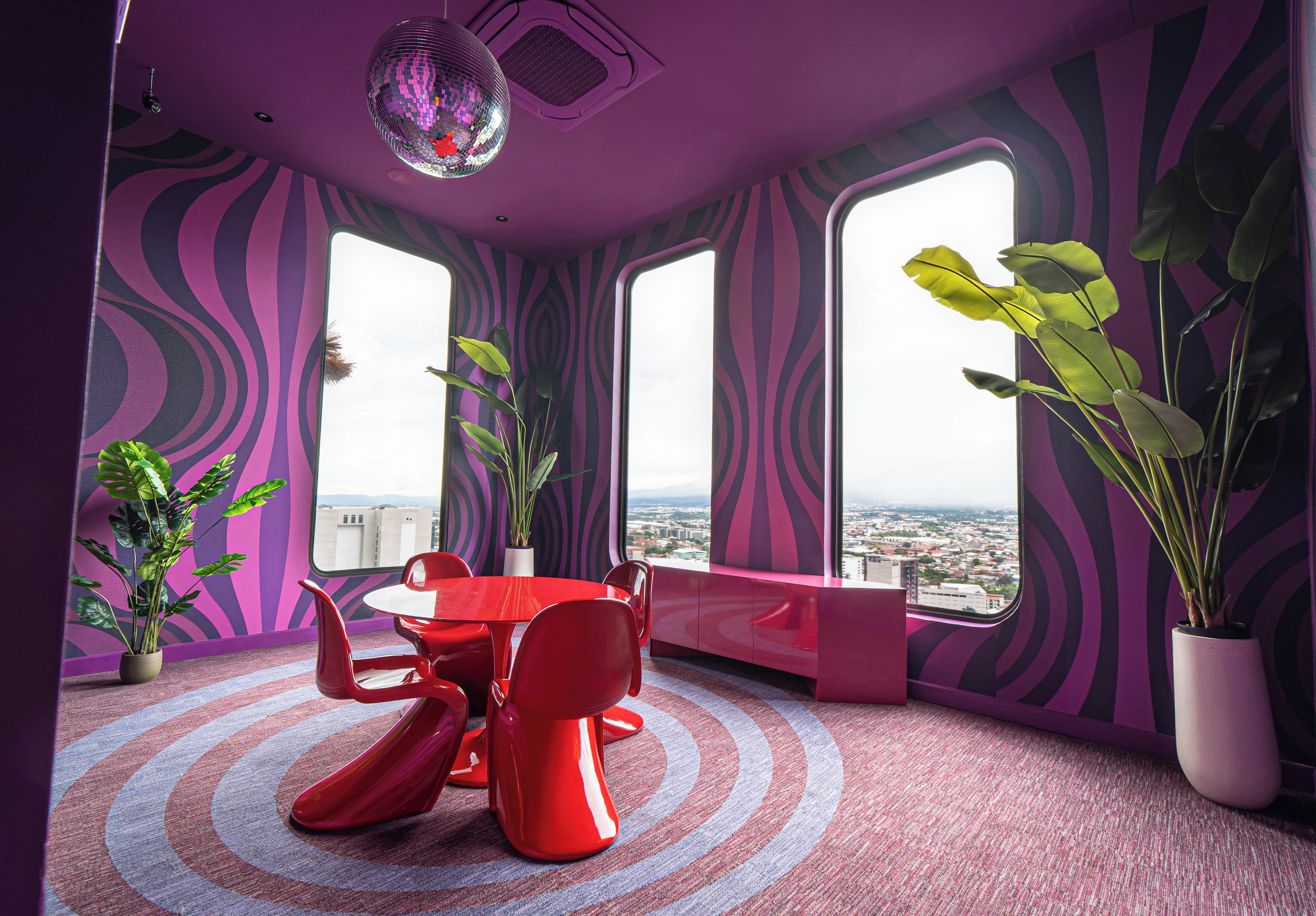 A room with a red table and chairs, large windows, and potted plants.