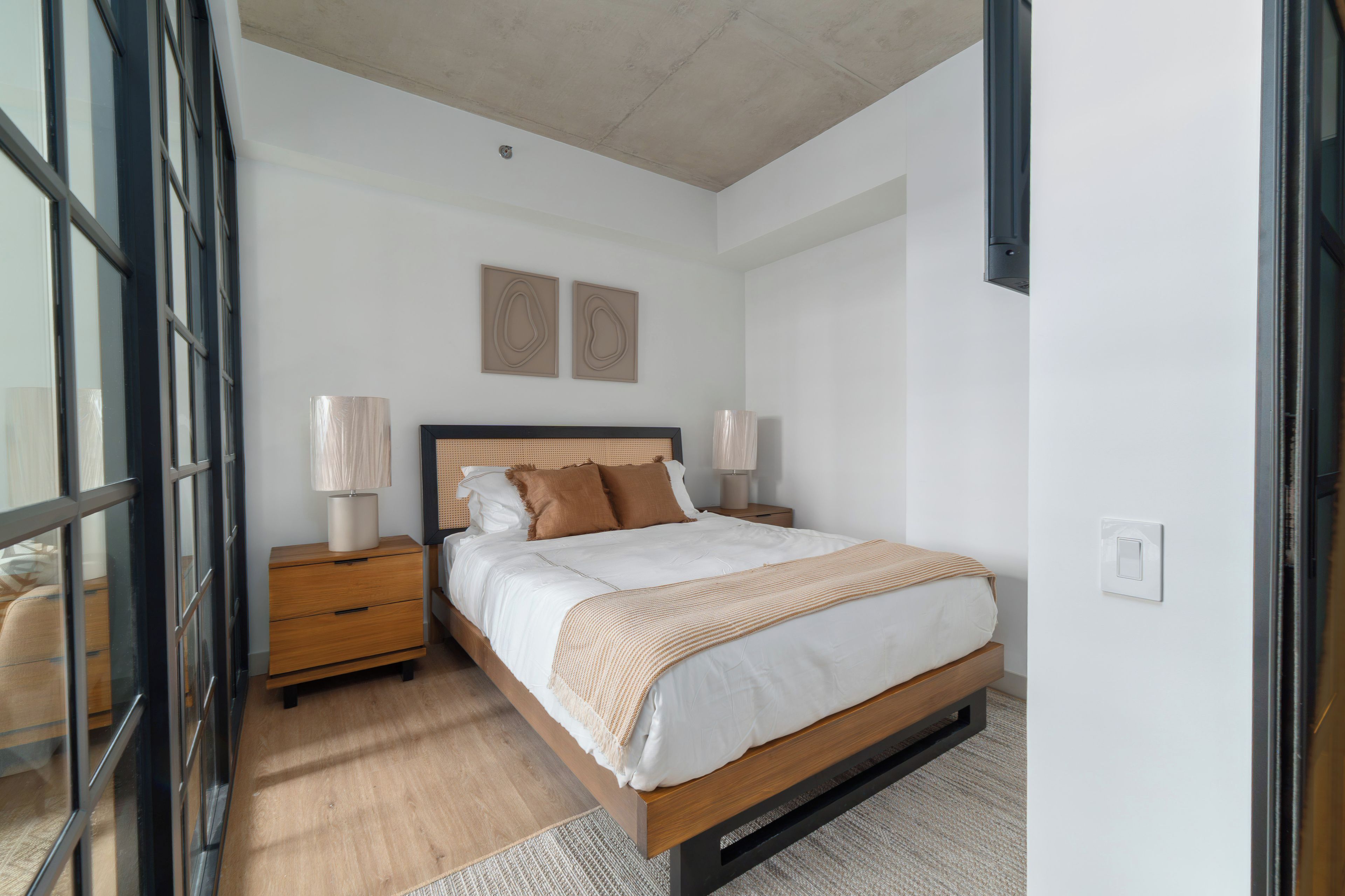 Un dormitorio con una cama de madera, dos mesitas de noche, una lámpara de noche y una ventana con vistas, además de dos lámparas fijadas a la pared.