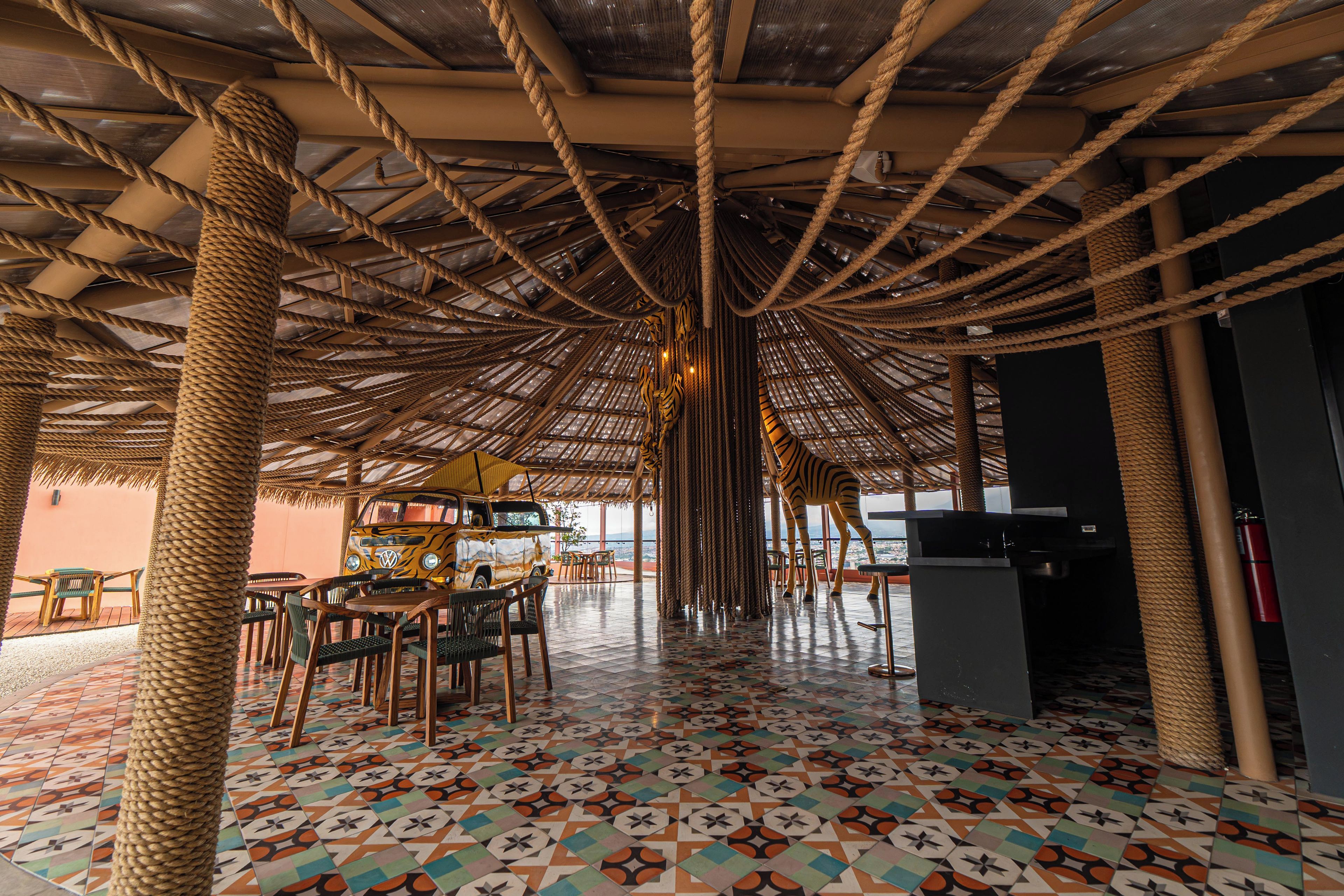 En inomhusrestaurang med ett halmtak, trämöbler och ett mönstrat golv.