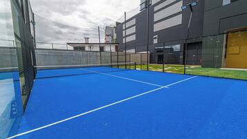 An outdoor tennis court with a net, surrounded by a fence and adjacent buildings.