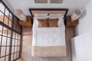 A neatly made bed with white linens and two brown pillows, a wooden headboard, and a woven rug at the foot of the bed.