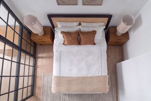 Una cama bien hecha con sábanas blancas y dos almohadas marrones, cabecera de madera y un tapete tejido al pie de la cama.