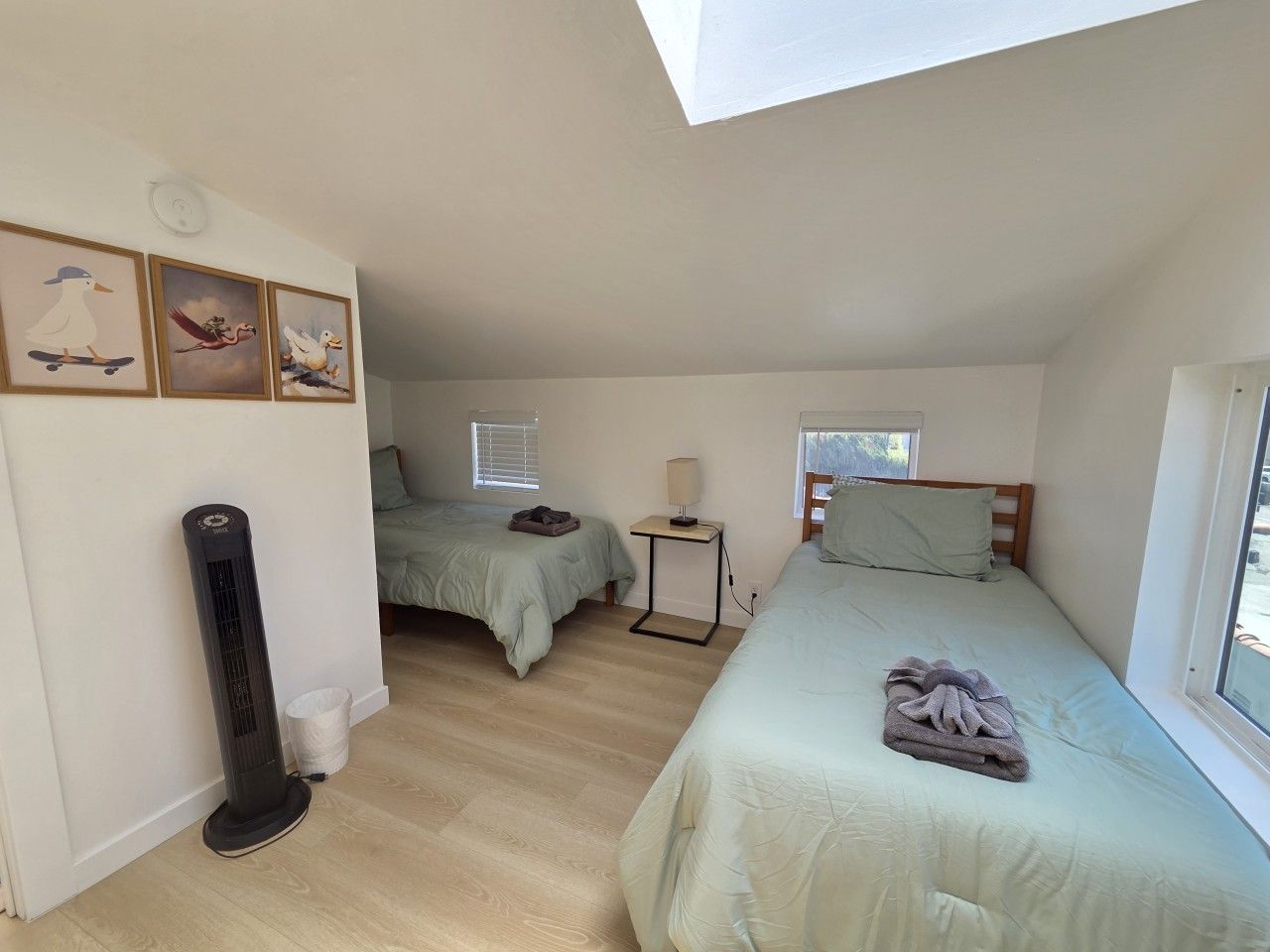 Une chambre avec deux lits, un radiateur mobile, une petite table avec une lampe et une fenêtre qui laisse entrer la lumière naturelle.