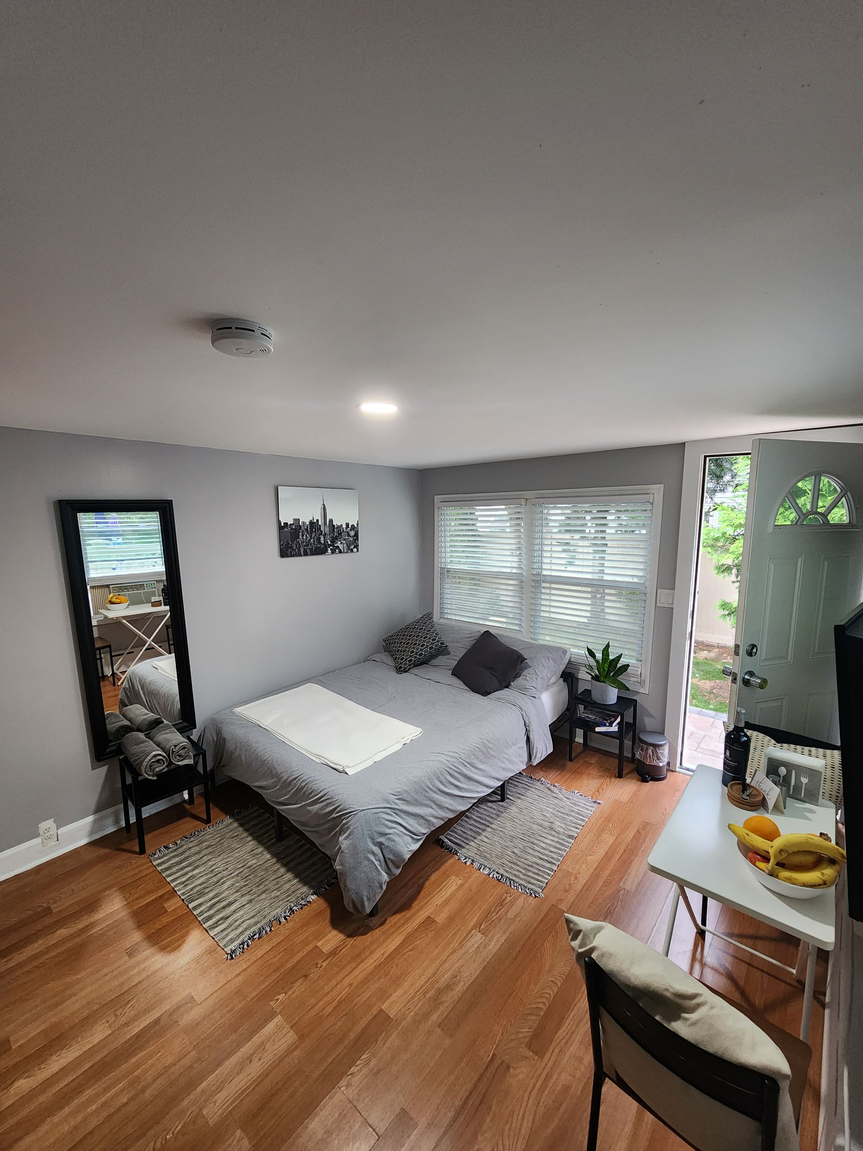 A bedroom with a bed, a mirror, a window with blinds, a small table with a plant, and a door leading to an outdoor area.