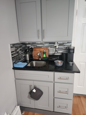 A kitchen with grey cabinets, a black countertop, a sink, a cutting board, a kettle, and a coffee maker. - Cozy studio in a lovely Forest Hills neighborhood (Forest Hills)