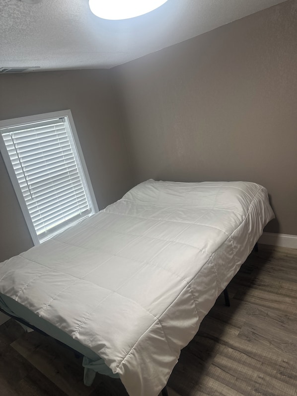 A small, single-bed room with a white bedspread, a window with blinds, and a wooden floor.