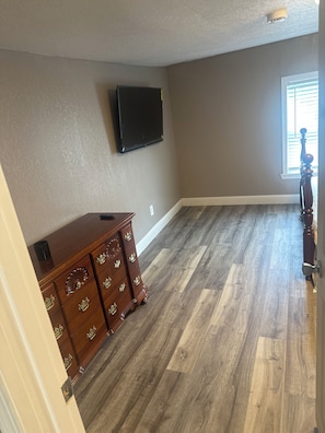 A room with a wooden dresser, a flat-screen TV mounted on the wall, and a window with blinds.