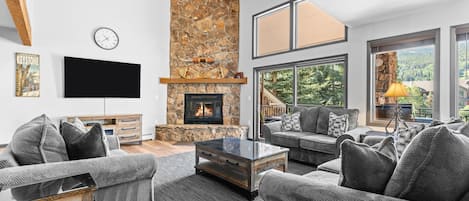 A modern living room with a stone fireplace, comfortable seating, a flat-screen TV, and large windows offering a view of the outdoors.