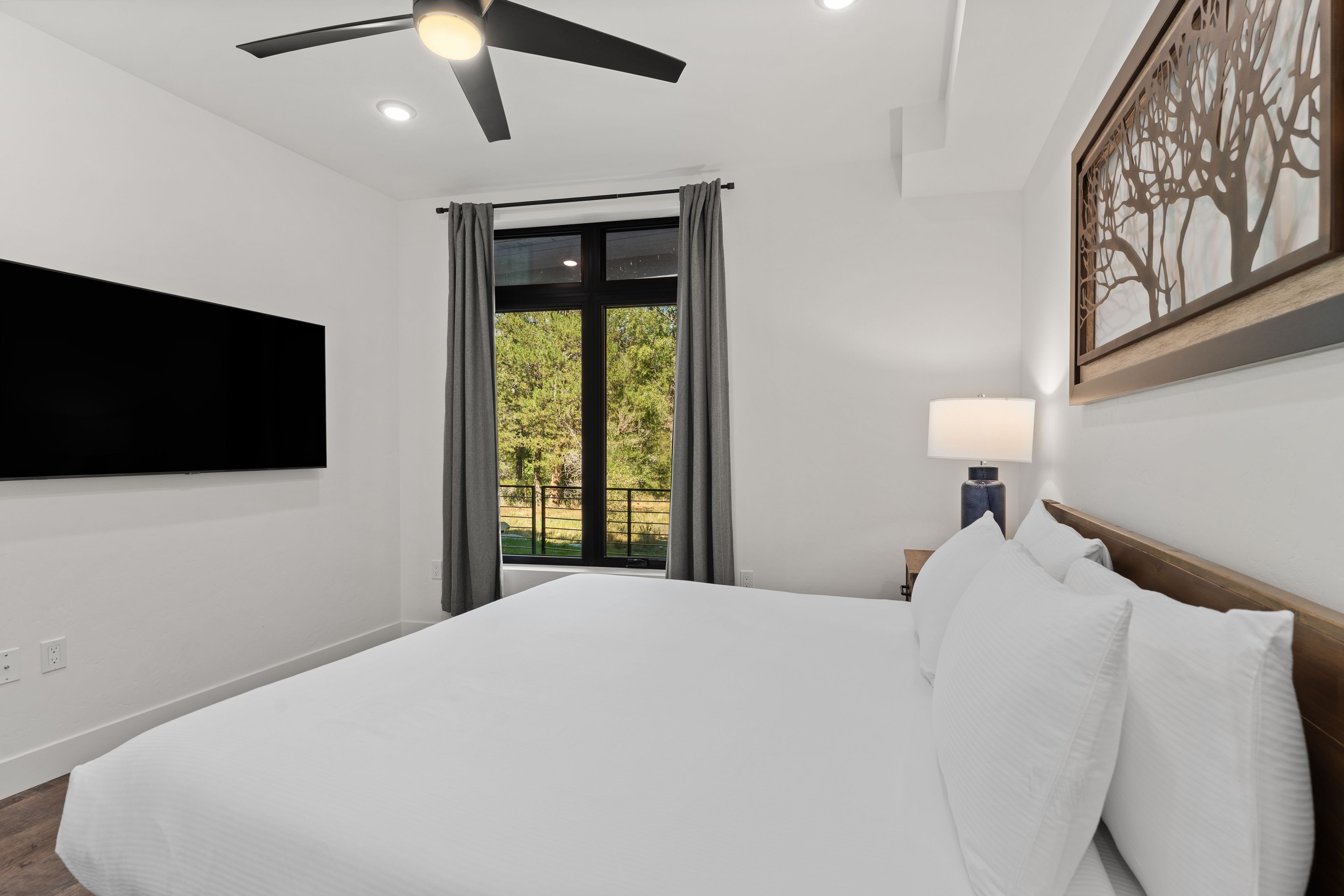 A hotel room with a bed, a flat-screen TV mounted on the wall, a ceiling fan, and a window with curtains.