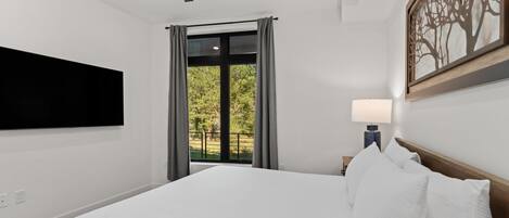 A hotel room with a bed, a flat-screen TV mounted on the wall, a ceiling fan, and a window with curtains.