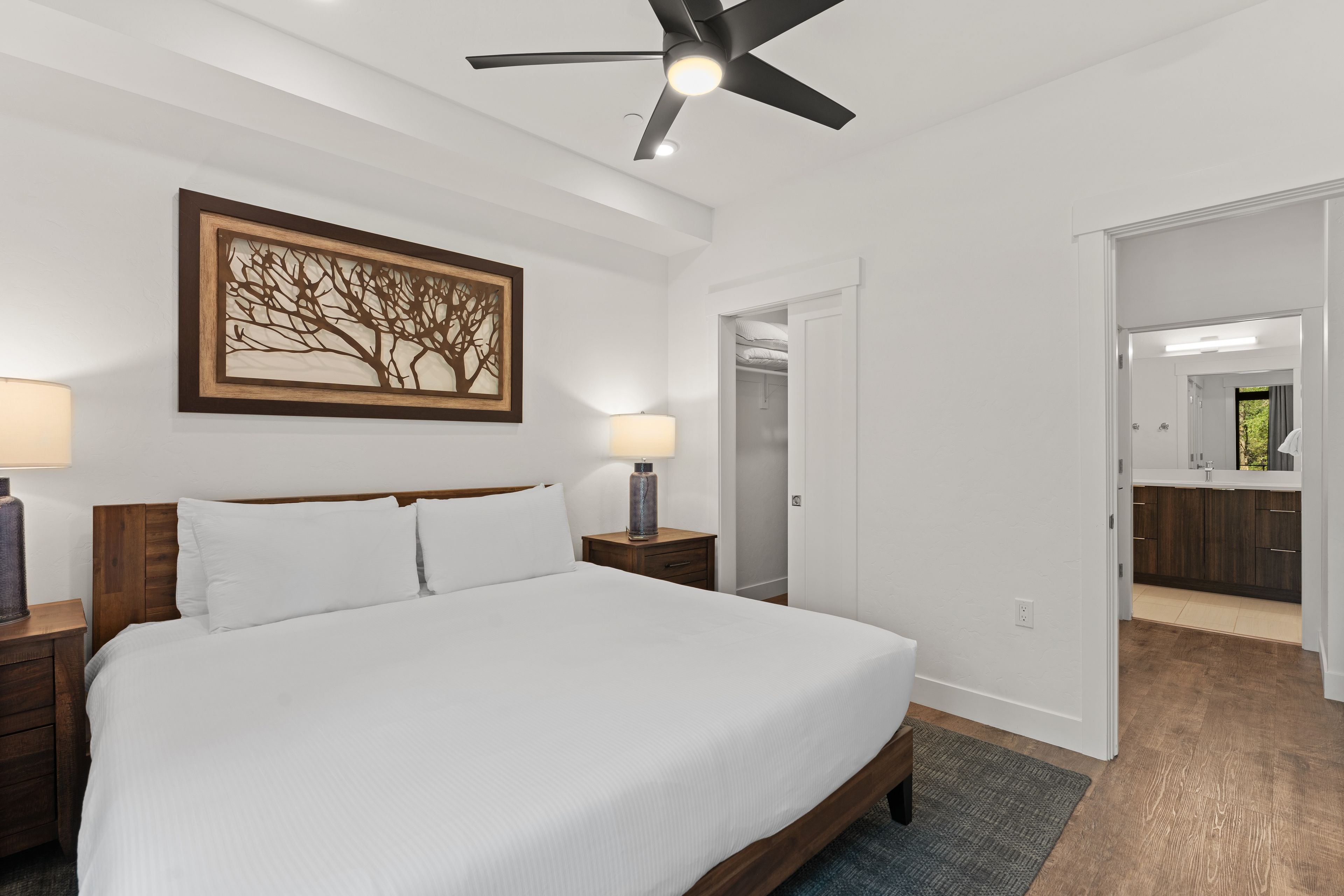 A bedroom with a bed, nightstands, a ceiling fan, a wall-mounted artwork, and a closet with a mirror.