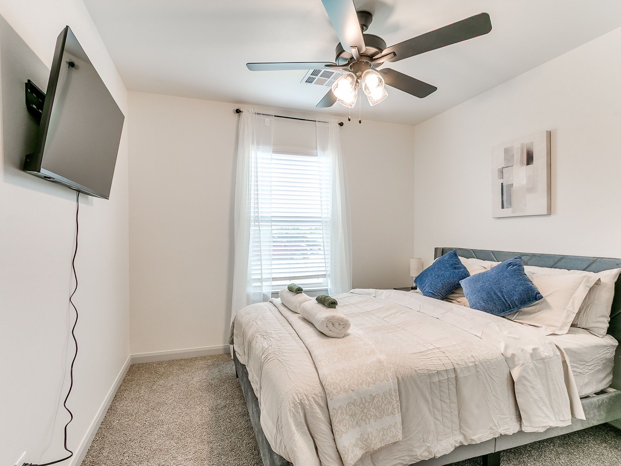 A bedroom with a bed, a ceiling fan, a TV mounted on the wall, and a window with blinds.