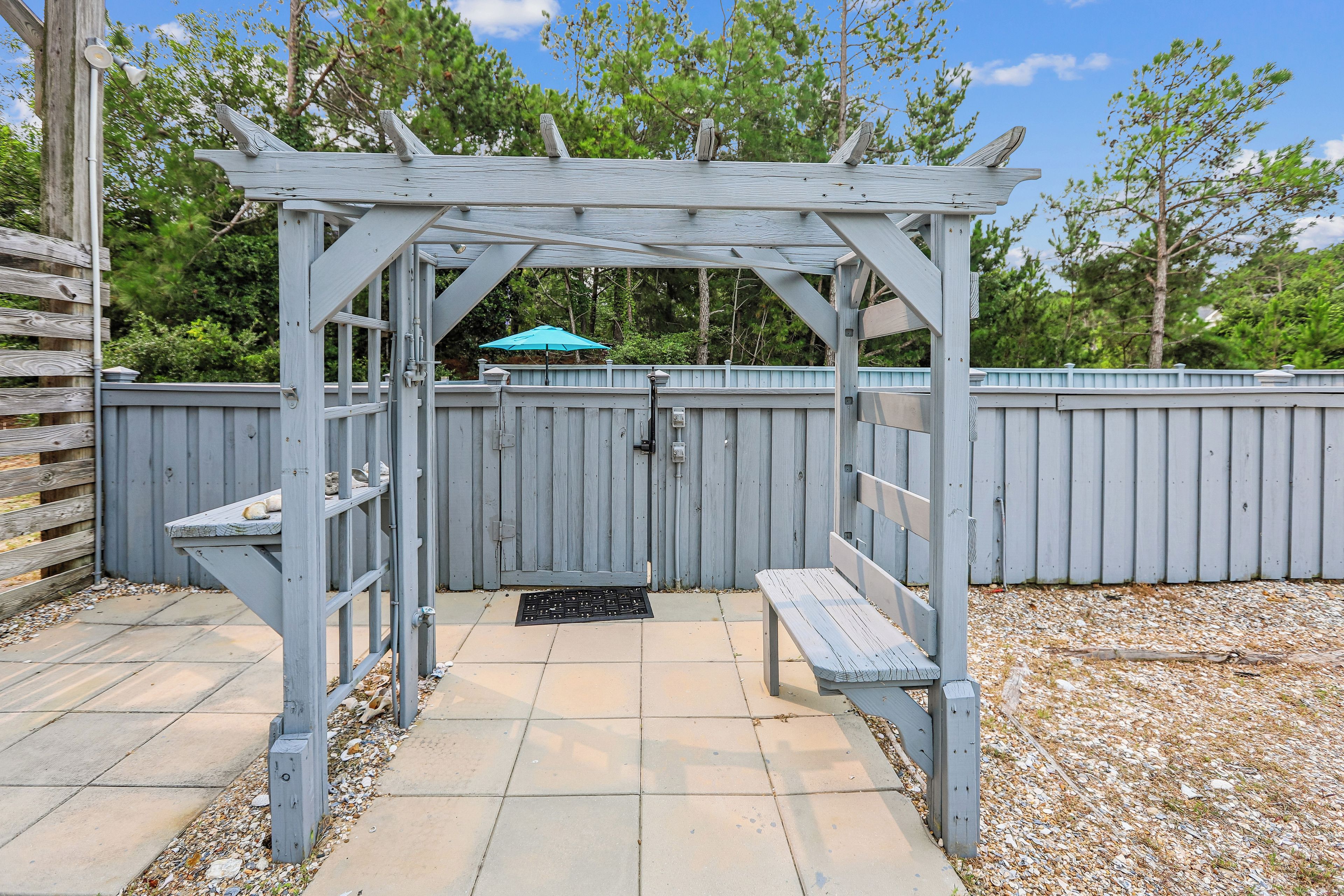 A wooden pergola with a bench and a door, surrounded by a wooden fence and a blue umbrella.