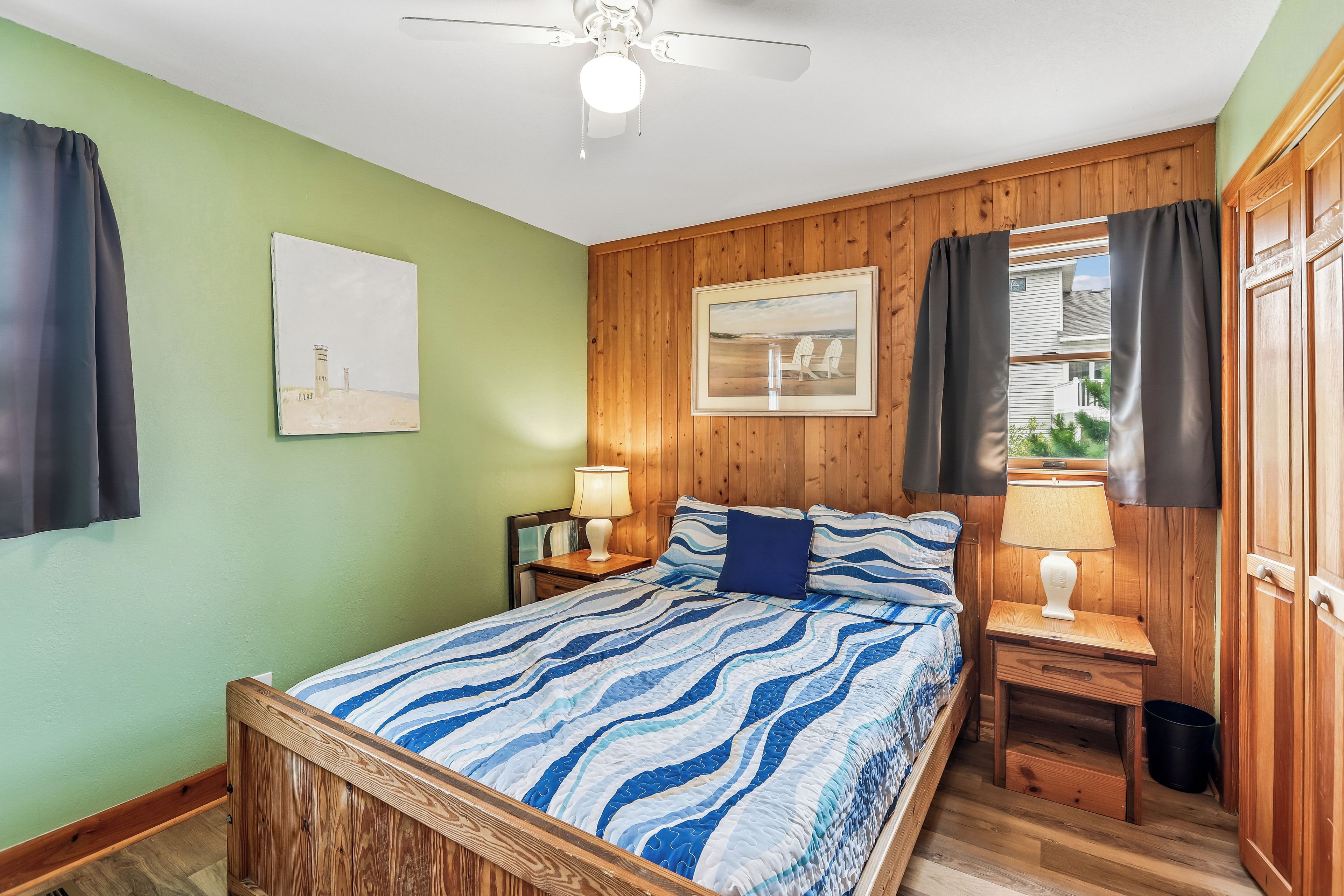 Une chambre à coucher comprenant un lit, une table de chevet, une lampe, un ventilateur de plafond, un tableau encadré et une fenêtre avec des rideaux.