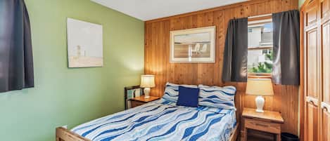 Un dormitorio con cama, mesita de noche, lámpara, ventilador de techo, cuadro enmarcado y ventana con cortinas.