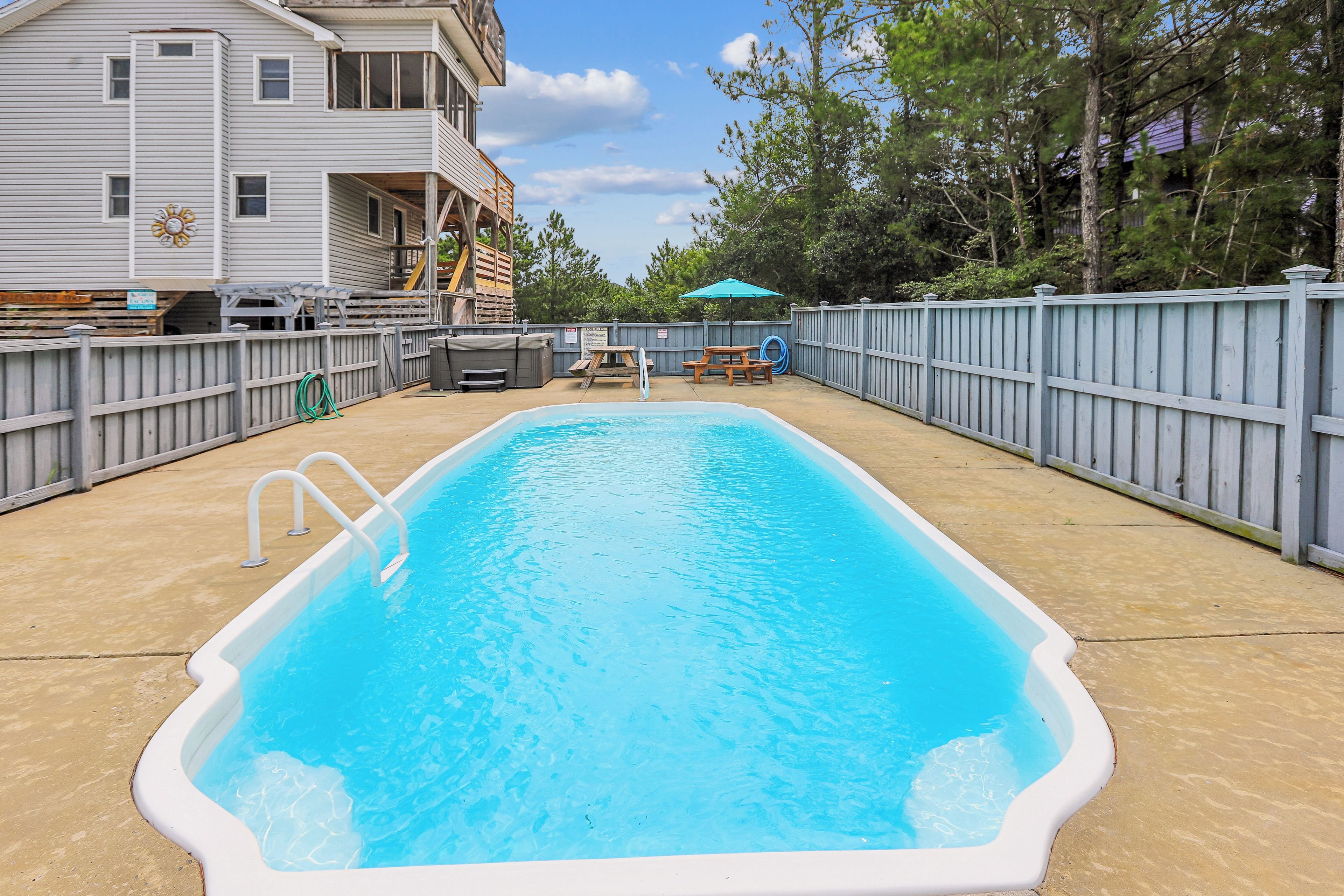 A rectangular swimming pool with a ladder on one side, surrounded by a wooden fence, with a picnic table and umbrella in the background.