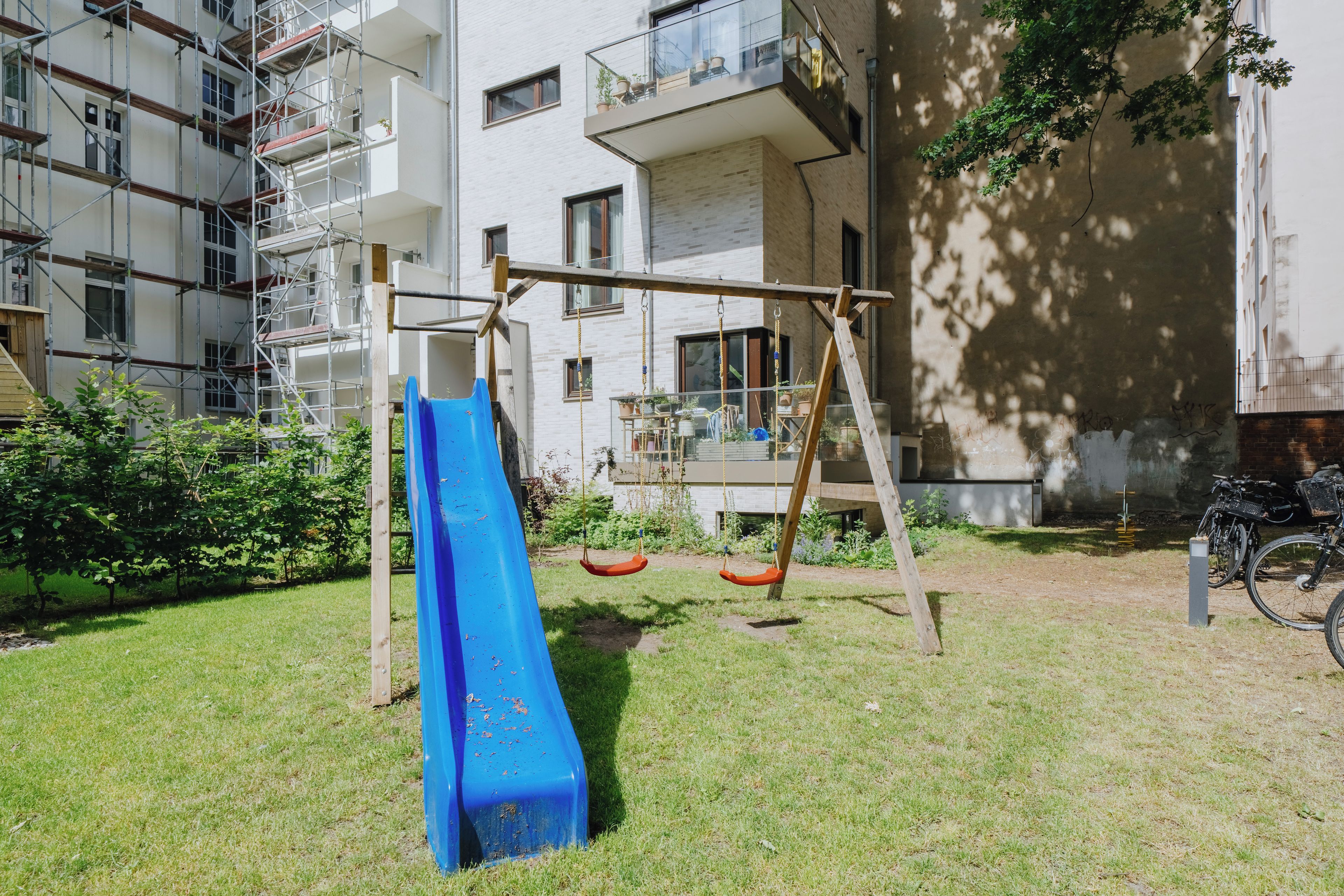 Ein Spielplatz mit einer blauen Rutsche und Schaukeln, umgeben von einer Rasenfläche und einem Gebäude mit Gerüst.
