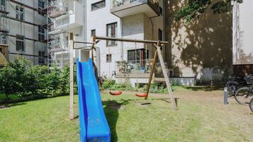 Ein Spielplatz mit einer blauen Rutsche und Schaukeln, umgeben von einer Rasenfläche und einem Gebäude mit Gerüst.
