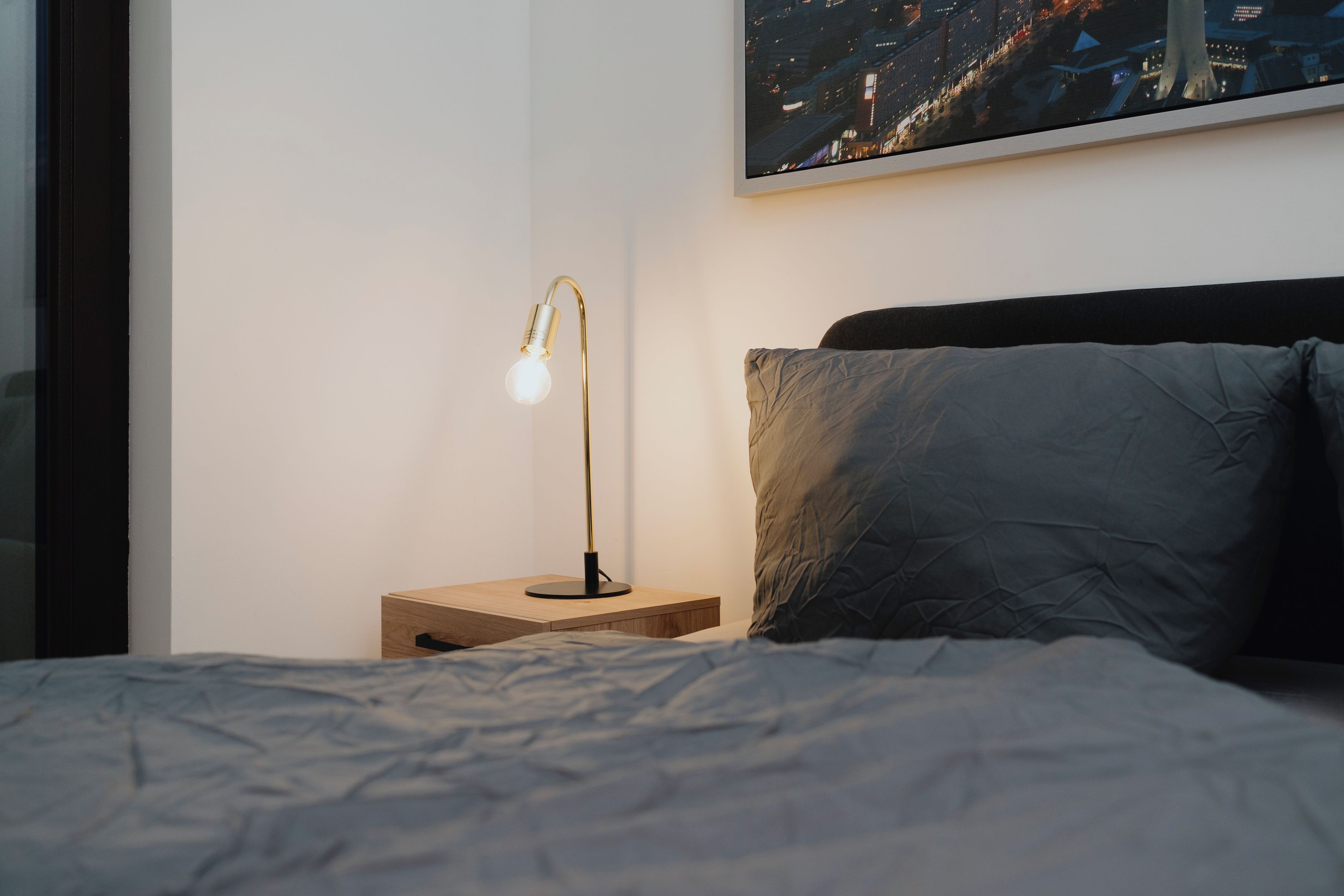 Ein ordentlich bezogenes Bett mit einer grauen Decke, ein Nachttisch aus Holz mit einer Lampe und ein Stadtbild an der Wand.