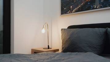 Ein ordentlich bezogenes Bett mit einer grauen Decke, ein Nachttisch aus Holz mit einer Lampe und ein Stadtbild an der Wand.
