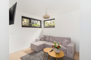 Ein modernes Wohnzimmer mit einem grauen Sofa, einem kleinen runden Tisch mit Blumenvase und einem an der Wand befestigten Fernseher.