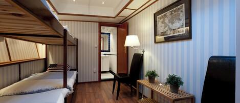 Une chambre d’hôtel avec un lit superposé, un bureau avec une lampe, une chaise, une petite table avec une plante et une carte du monde encadrée accrochée au mur.
