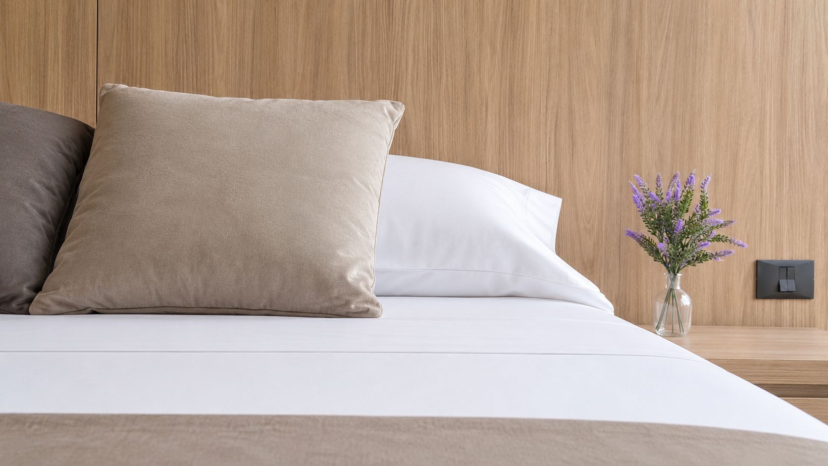 Cama com travesseiro bege e um travesseiro branco, um vaso com flores roxas em uma mesa de cabeceira e cabeceira de madeira.
