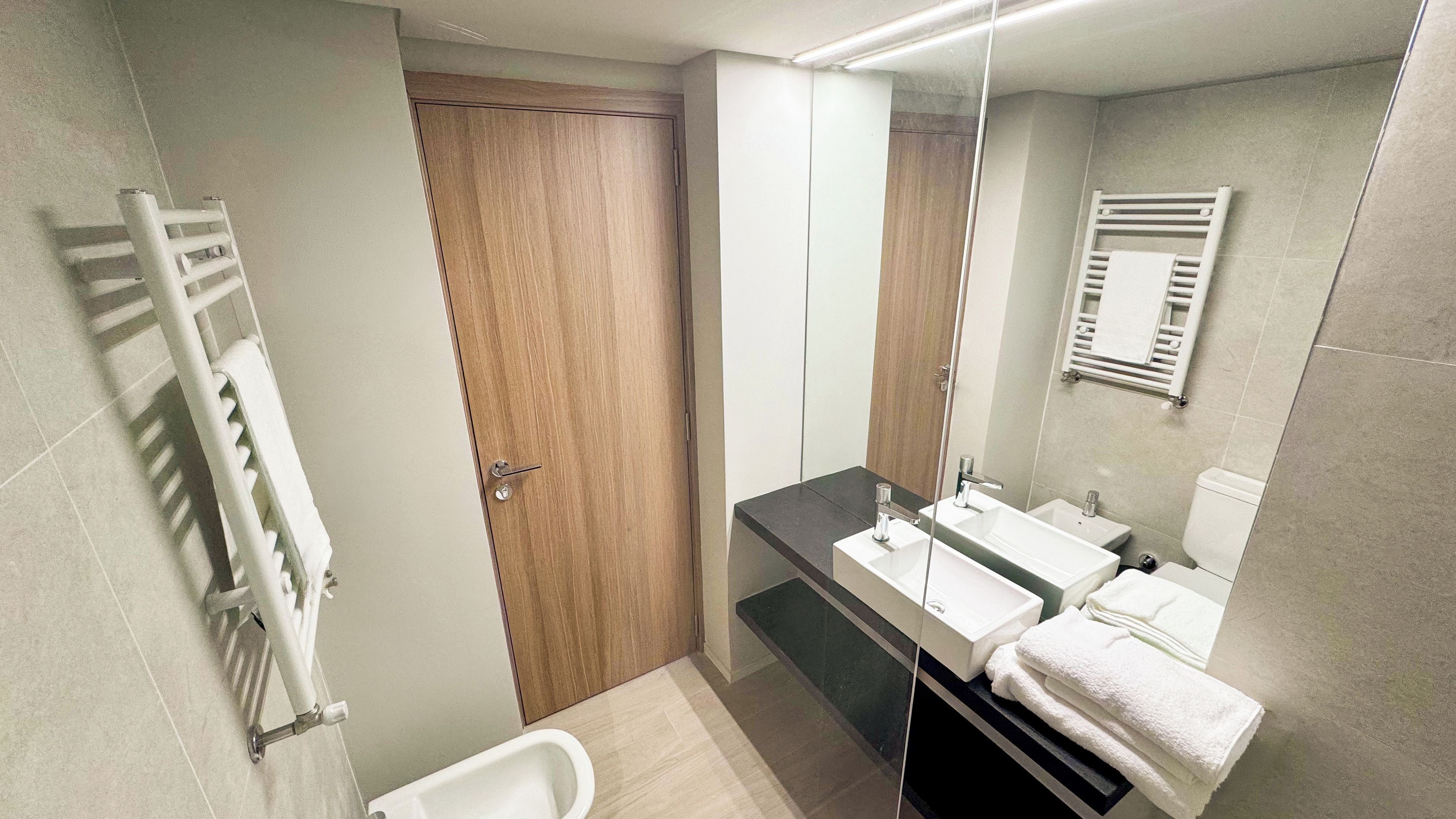 A modern bathroom with a wooden door, a glass shower enclosure, a white bathtub, and a vanity with two sinks and a mirror.