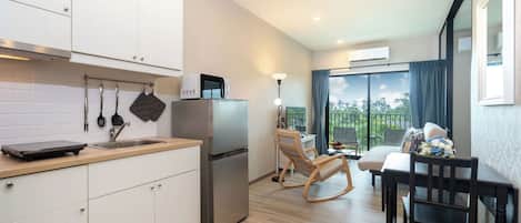 Une cuisine compacte avec des meubles blancs, un four à micro-ondes encastré et un réfrigérateur. On y trouve un coin repas avec une table et des chaises en bois, ainsi qu’un balcon offrant une vue sur la verdure.
