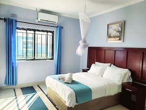 Een hotelkamer met een bed, nachtkastje, raam met gordijnen, airconditioning en een ingelijst schilderij aan de muur.