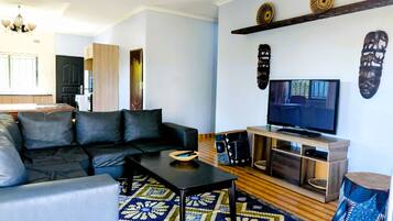 Un salon avec un canapé noir, une table basse, un téléviseur posé sur un meuble en bois, des tapis à motifs et des œuvres d’art décoratives accrochées au mur.