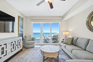A spacious living room with a large TV, a round coffee table, and a sofa. There is a balcony with a view of the ocean. - Steps to The Beach • Resort Pools • Pier Park (Panama City Beach)