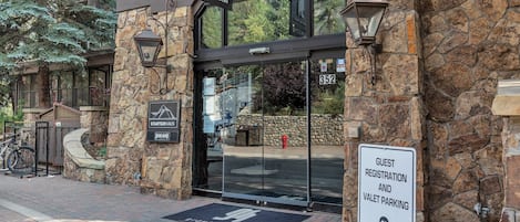 Entrada de un edificio con letrero para registro de huéspedes y servicio de valet, fachada de piedra y felpudo con un logotipo.