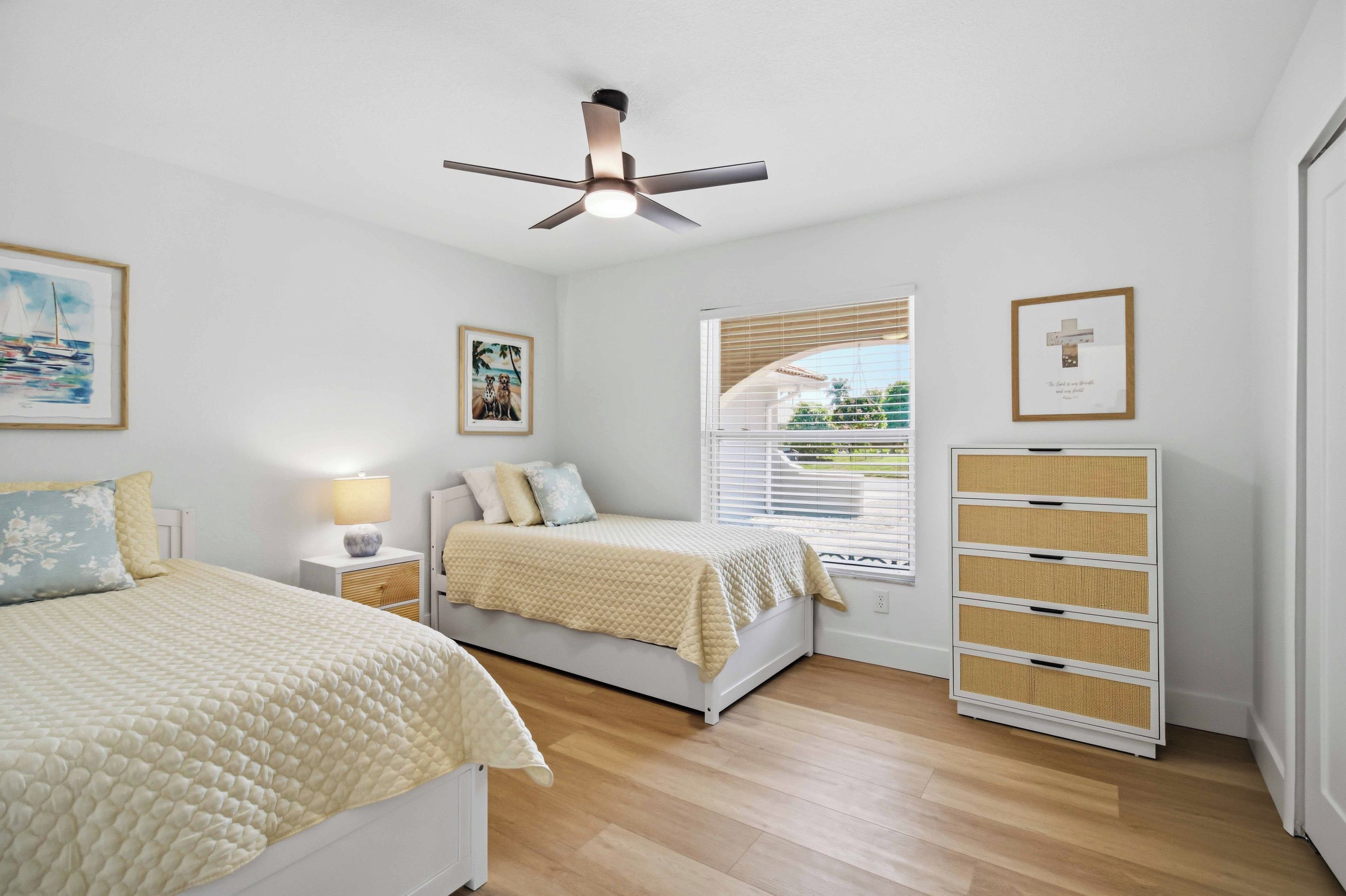 A bedroom with two beds, a ceiling fan, a window with blinds, and a bedside table with a lamp.